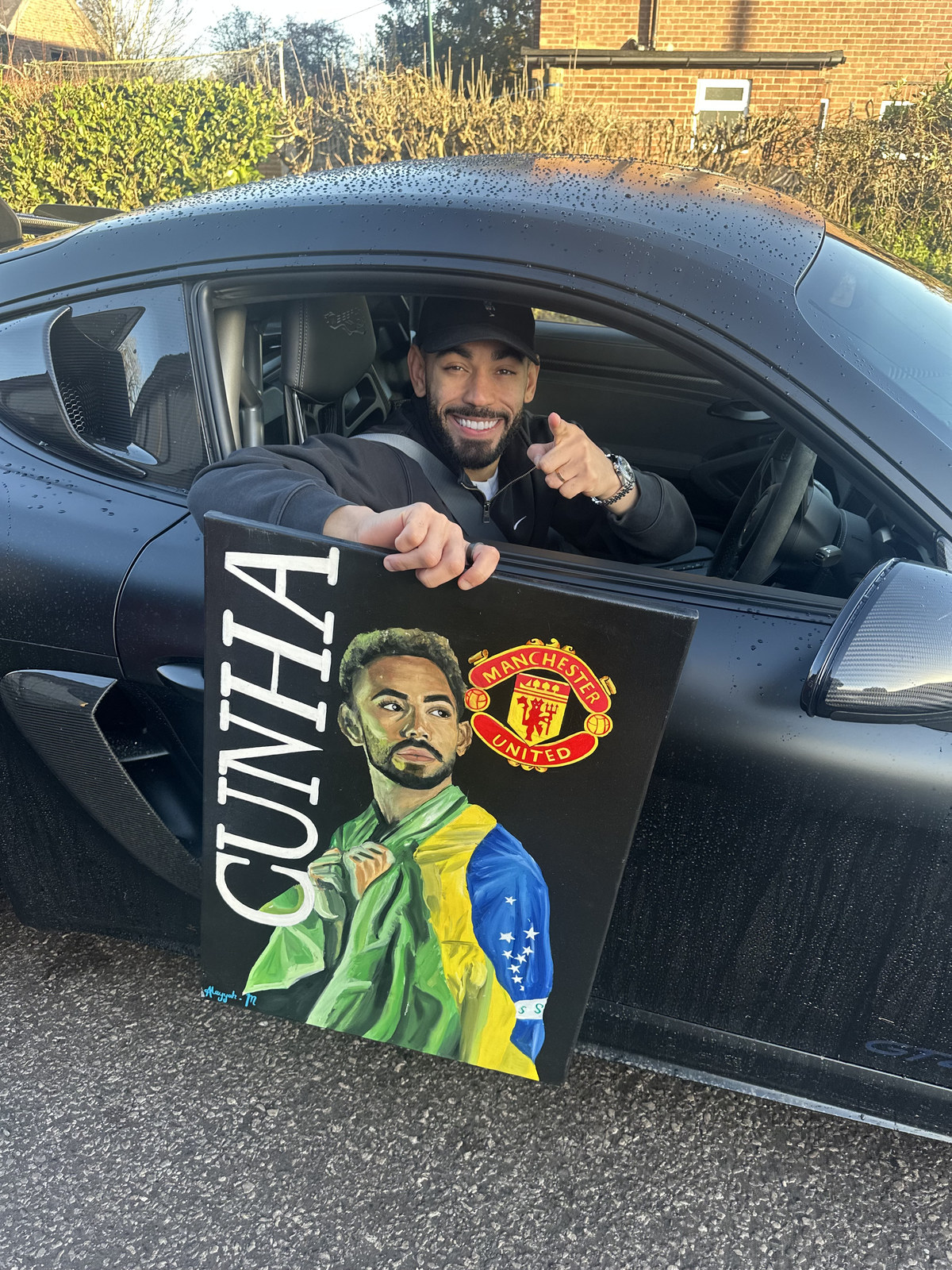 Footballer Matheus Cunha smiling from a black sports car while holding his painted portrait